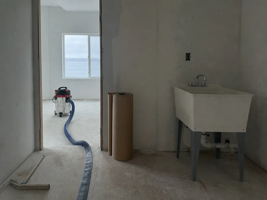 A wet vacuum with a lay flat discharge hose run from a doorway to a utility sink, a squeegee and a roll of floor protection beside the threshold