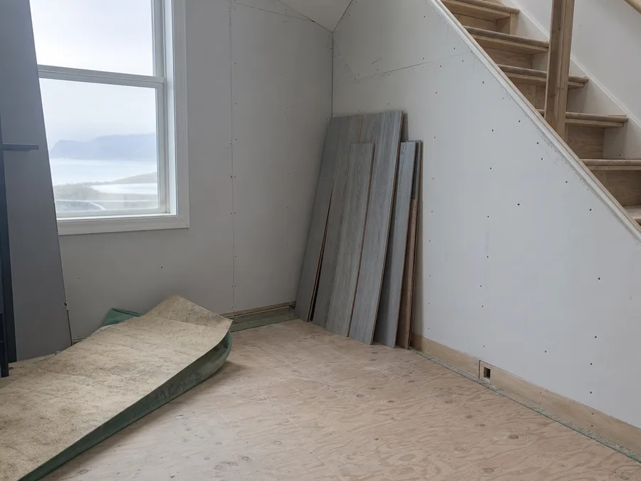 A lower level room with a run of laminate lifted and stacked against the wall, the underlay peeled back off the subfloor beside a boxed in staircase