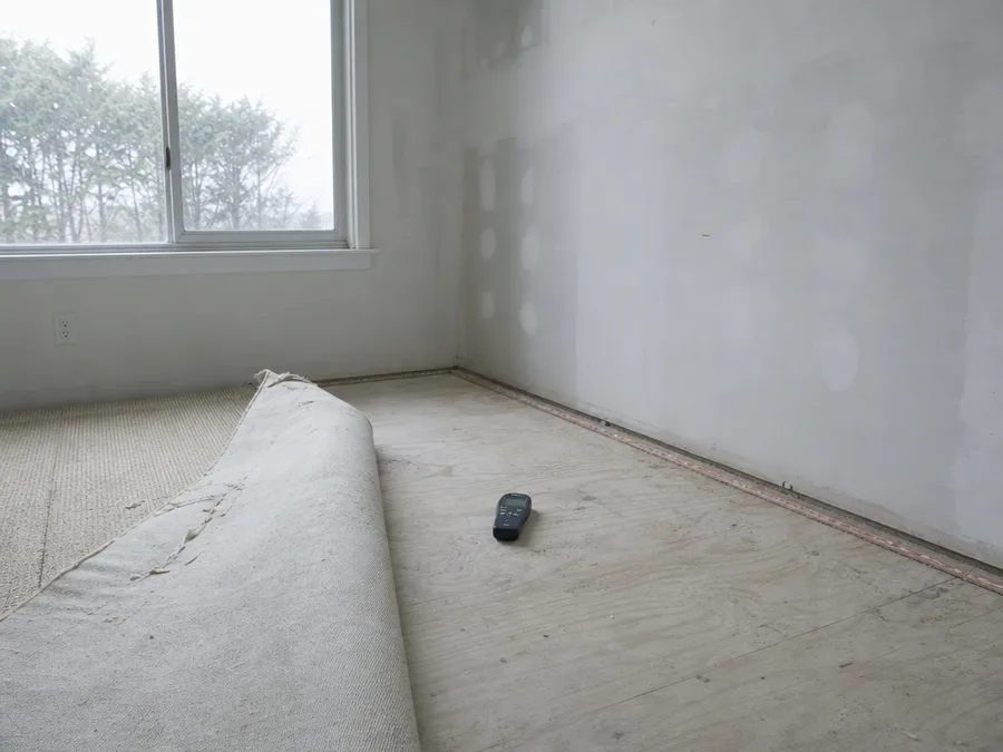 A folded corner of carpet pulled back from a tack strip with a moisture meter resting on the exposed subfloor