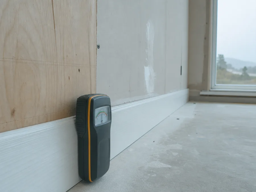 A moisture meter standing against a painted baseboard beside a part boarded wall, the dial on its face and the grain of the plywood behind both in focus