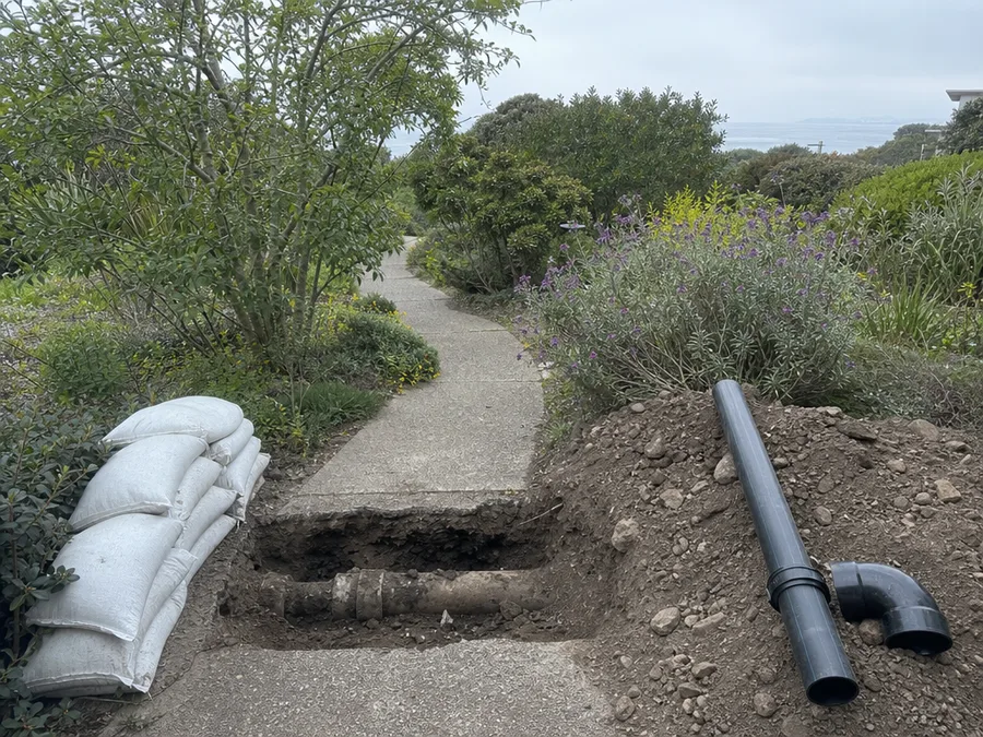 An open trench cut across a front garden path with the old sewer lateral exposed in the bottom of it, a length of new black pipe and an elbow fitting laid on the spoil heap alongside and sandbags stacked at the edge